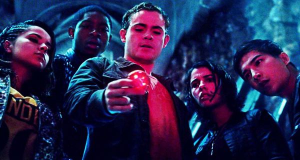 The Power Rangers cast, slightly damp, looking at a glowing red object that casts their faces in red. From left to right we have the Yellow Ranger, the Blue Ranger, the Red Ranger (who is holding the glowing red coin), the Pink Ranger and the Black Ranger. They all look on with expressions of surprise. This is a still from the 2017 Power Rangers movie.