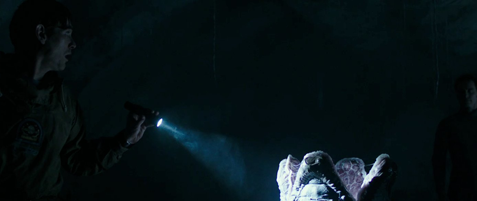 A man with a flashlight points it towards an Alien egg, with another man in the background nearly invisible in the darkness. This is a still from the film Alien Covenant