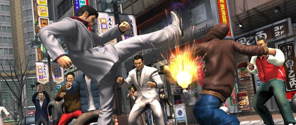 A man in a white suit is kicking another man in a brown bomber jacket in front of a crowd of people. This is a still from the game Yakuza