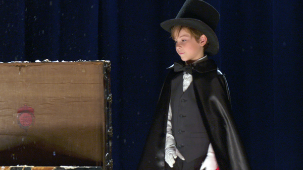 Jacob Tremblay as Peter, dressed up as a pint sized magician in black top hat and cape, standing next to an open trunk on the floor on a well lit stage. This is a still from the movie Book of Henry