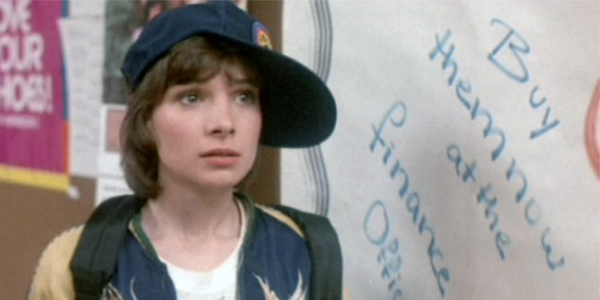 Paul, with her Nostromo ballcap tilted slightly askew, stands in front of a white board that reads "buy them now from the finance office." This is a still from the film Neon Maniacs