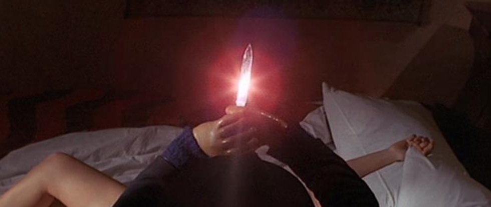 a mans hands hold a knife aloft, the blade lit red in a lens flare. Below him on a bed stretches the naked limbs of a woman. This is a still from the film The Black Belly of the Tarantula.