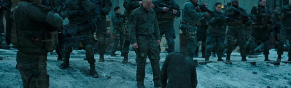Caesar, the ape, sitting on his knees in the snow in a supine position to a row of soldiers. This is a still from the film War for the Planet of the Apes.
