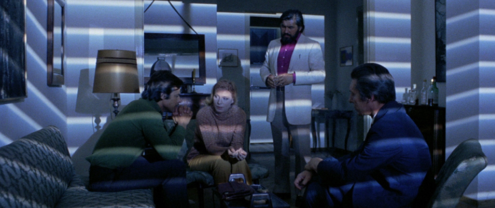 A group of people in a room, looking somber the light across their faces showcasing the lines of a set of near drawn blinds. This is a still from Short Night of Glass Dolls.