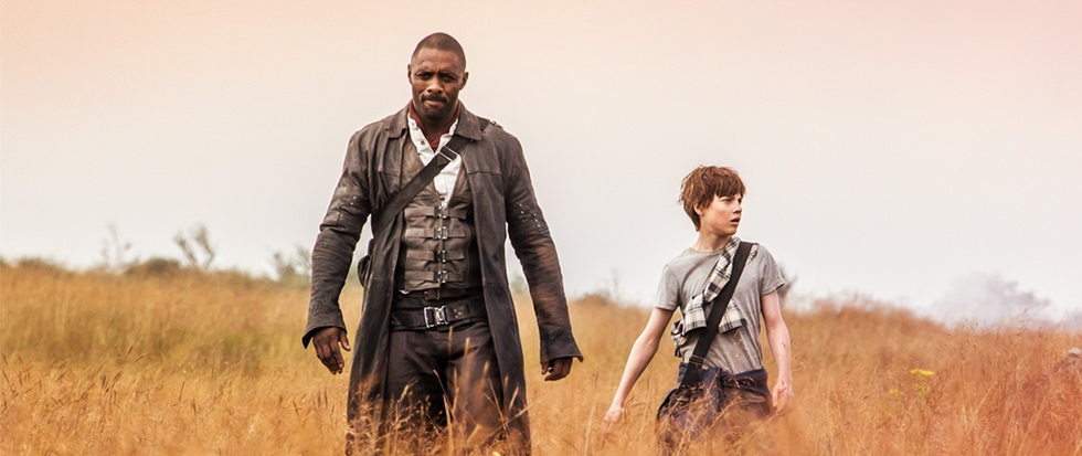 Idris Elba in a long coat and a shoulder holster, walking with a young boy through a yellow brown field. This is a still from the movie the Dark Tower.