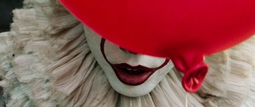 Red lips in a white face with a ruffle behind, a red balloon partially consider the face. This is a still of Pennywise from It.
