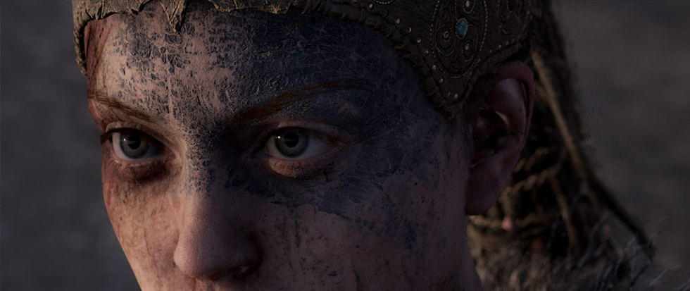 A womans eyes peer out from a blue painted face, her hair running in locs behind her head. This is a still from Hellblade; Senua's Sacrifice