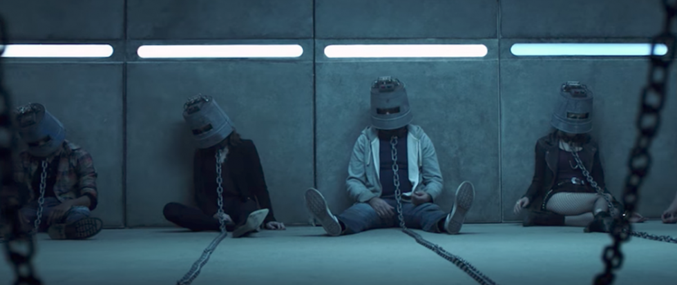 Foru figures sitting on the ground with buckets on their head, chains attaached to the adjoining wall. This is a still from the horror film Jigsaw.