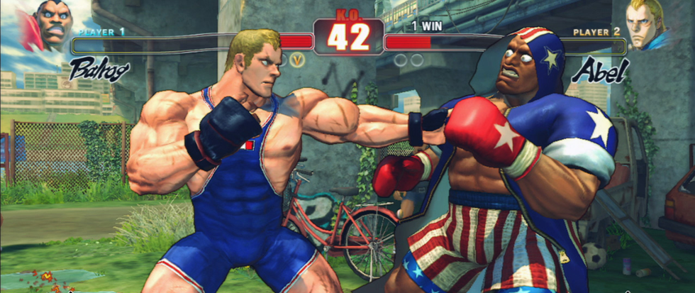 A blonde man in a blue one piece leotard punches out with a boxing fist towards a dark skinned man in an American flag boxing outfit.