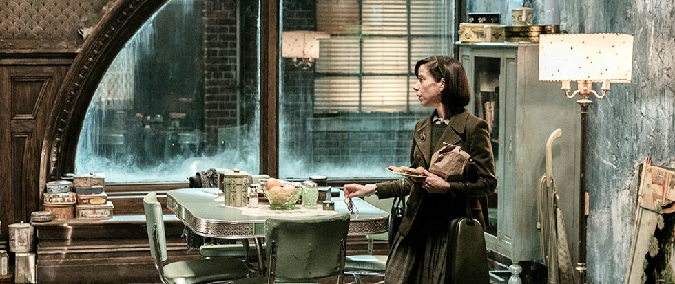 Sally Hawkins standing as her character in Shape of Water in a 1950's dark blue green accented kitchen a large window open in the background.