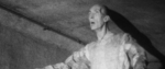 In grainy black and white, the top half of a very tall man with his arms outstretched and his mouth open.