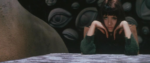 A Japanese woman with her head in her hands and two miscolored eyes sitting at a table in front of a wall of sculptural eyes.