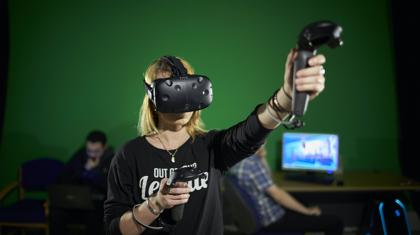 A young woman wears a virtual reality headset while holding a VR controller in each hand.
