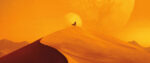 A tiny figure in a flowing cloak stands atop an enormous sand dune, the sun setting directly behind them.