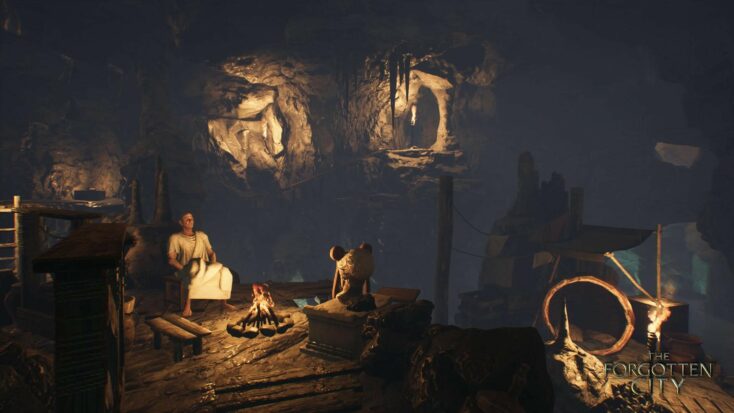 A man in a toga sits in a darkened cave next to a small campfire.