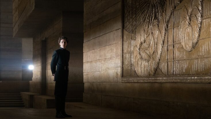 A young man in dark, well-tailored clothes stands stoically next to an elaborate stone freize.