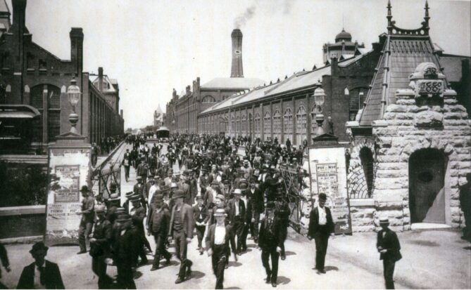 An old photograph showing a throng of people walking through the gates of a factory building.