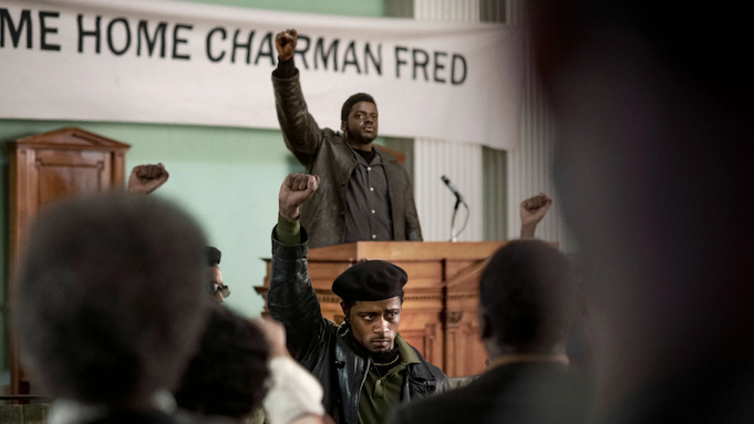 Fred Hampton (played by Daniel Kaluuya) stands at lectern with his fist raised in the air. Bill O’Neal (played by Lakeith Stanfield) stands on the floor directly in front of the lectern, raising a fist in an identical salute.