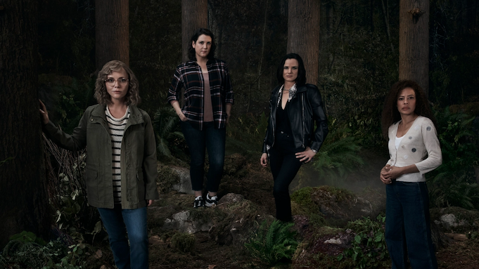 Four women, dressed in clothes ranging from high to low fashion, stand in a dark wood.