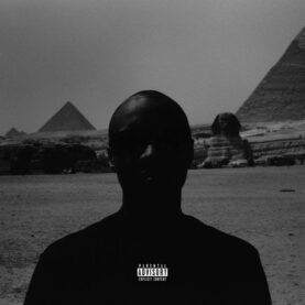 A photograph of a man standing in front the pyramids in Giza. The exposure is such that the subject is very dark, as if his features are totally obscured by shadow.