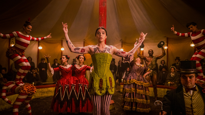 A tattooed woman in a corset and striped tights stands in a circus tent surrounded by acrobats, strongmen, and fire breathers.