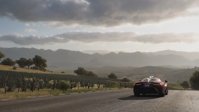 A souped-up sportscar drives on a deserted road through farmland. The sun sets behind a mountain range stretching before it.