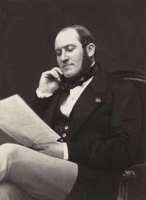 A photograph of Baron Haussmann. He wears dark mutton chops and sits reading a large document in a well-tailored suit.