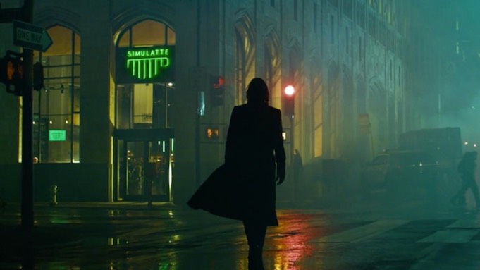 A man in a long coat walks rain-slicked city streets at night. In the distance a coffee shop's neon sign glows green.