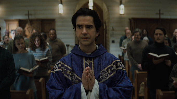 A priest in a deep purple chasuble walks up a church aisle towards the pulpit, hands clasped together as in prayer. 