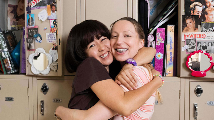 Two teenage girls (as played by 30-somethings) embrace enthusiastically in front of a bank of school lockers.