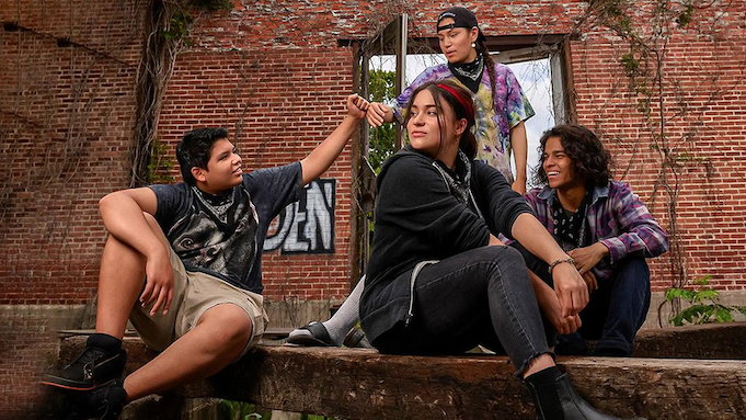 Four indigenous teenagers hang out on an abandoned lot, graffiti and vines covering the brick wall behind them.