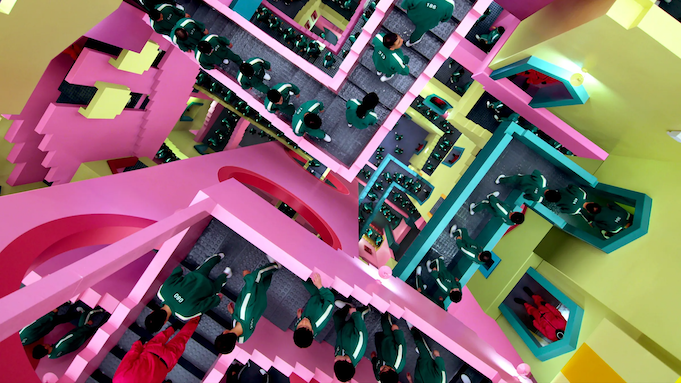 A top-down view of a maze of candy-colored stairways with many people dressed in matching green tracksuits climbing up and down, every which way.