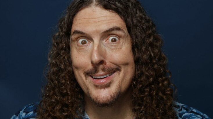 A close-up photograph of Weird Al Yankovic. He has long, curly brown hair and is making a goofy face with comically wide eyes and a crooked grin.