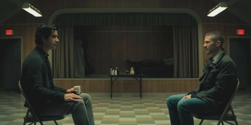 Two men sit in chairs facing each other in an empty auditorium. One of them is a priest. Both are dressed casually.