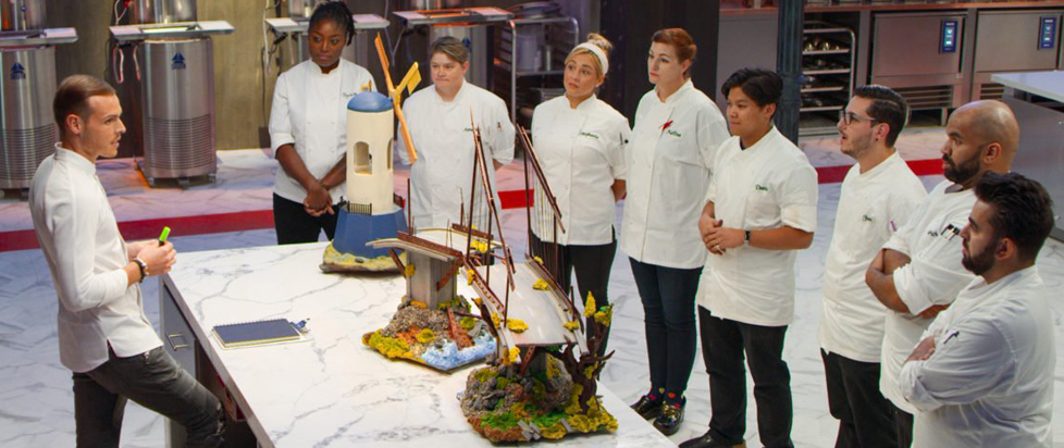 A master chocolatier stands behind a kitchen workstation lecturing to contestants on the reality show "School of Chocolate."