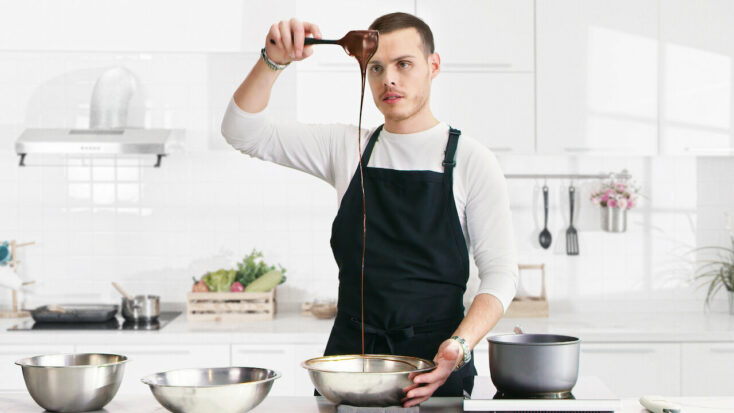A young man in an apron pours chocolate from a wooden spoon into a silver mixing bowl. He is examining the pour intently.
