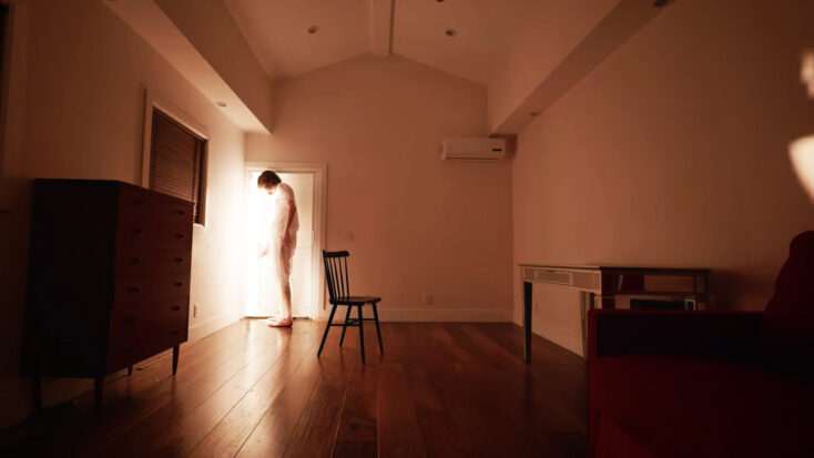 A scruffy ginger man in just his underwear tentatively opens a door from his small empty room to the outside.