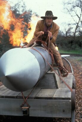A man in a leather duster and cowboy hat sits astride an enormous silver missile as flames erupt behind him.