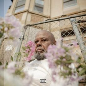 The cover of Saba's Few Good Things album featuring a stern-looking man sitting in a yard with a chain-link fence. Several delicate blooms are in the foreground, partially obscuring his face.
