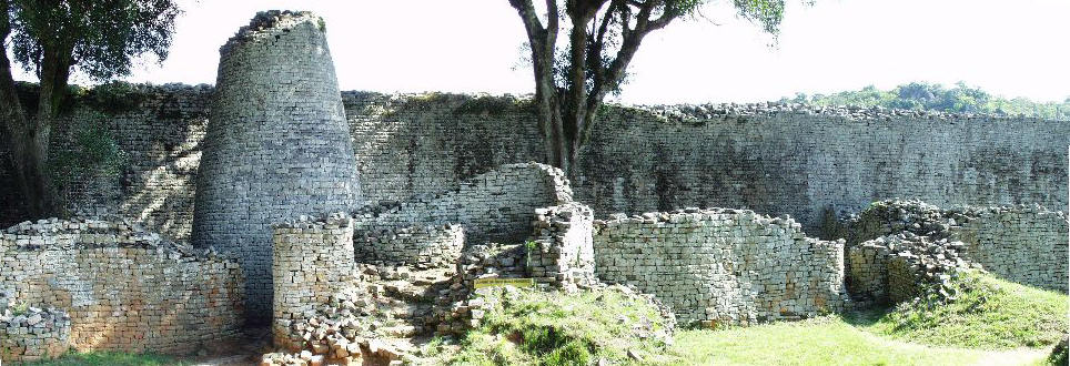 Stone ruins in the medieval city of Great Zimbabwe. Several low walls and a stone tower crumble into verdant green slopes.