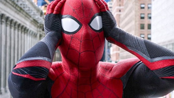 In a still from Spider-Man: No Way Home, Spider-man holds his hands to his head in a gesture of worry. Large city buildings stand tall behind him.