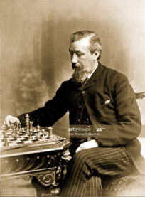 A sepia photograph of Joseph Henry Blackburne, the British chess player. He wears a dark suit and studies a chess board thoughtfully.