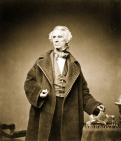 A sepia photograph of Samuel Morse. He is a white-haired man wearing an oversized topcoat and cravat.