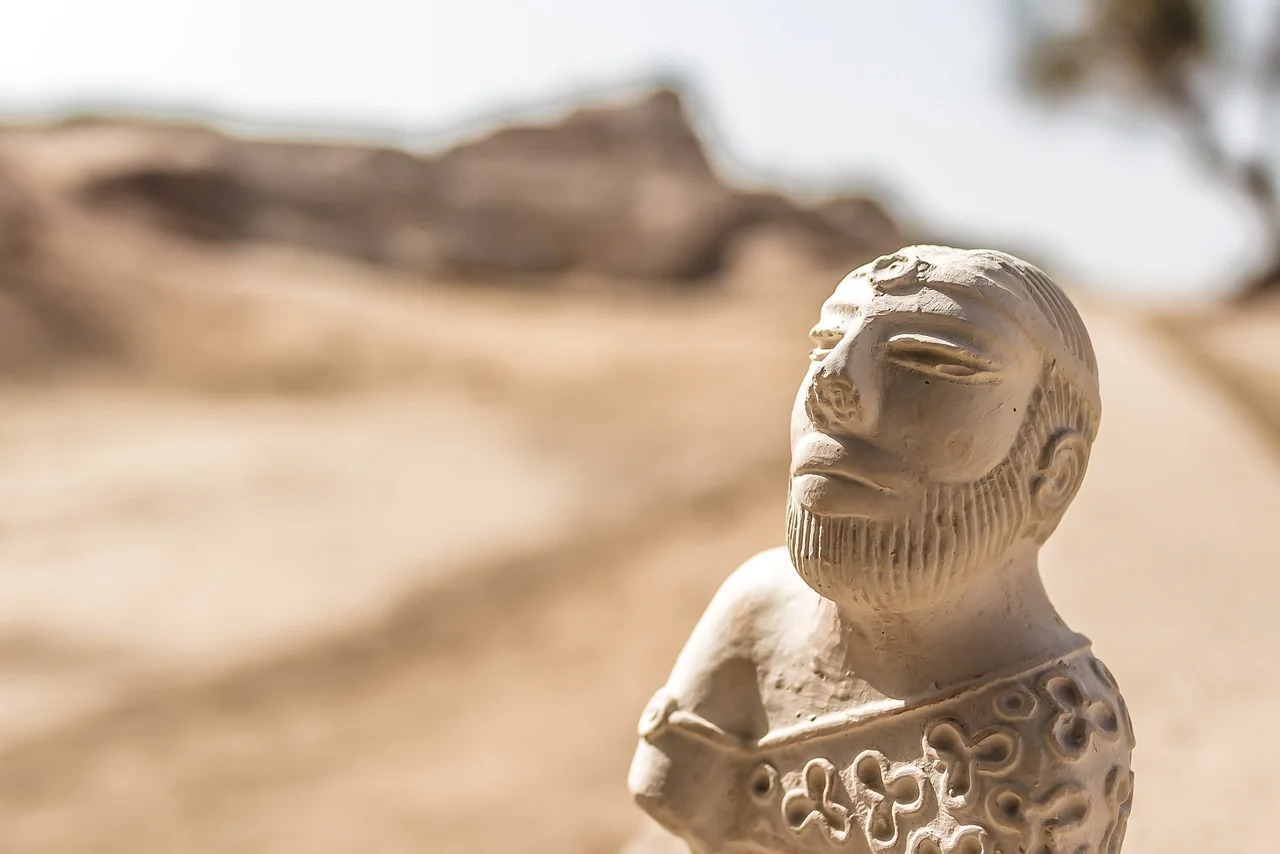 A clay statuette of a bearded man wearing an intricate floral garment sits in bright sun, framed by the ruins of Harappa in the distance.