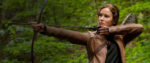 A still of Jennifer Lawrence as Katniss Everdeen in The Hunger Games, stands in thick woods and readies a bow and arrow to take a shot.
