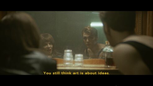 A screenshot from Immortality where two people stare into a mirror, one with short hair and full face of makeup, the other with longer hair and lipstick and leather, with a bottle of liquor and some glasses between them. The subtitle reads: "You still think art is about ideas."