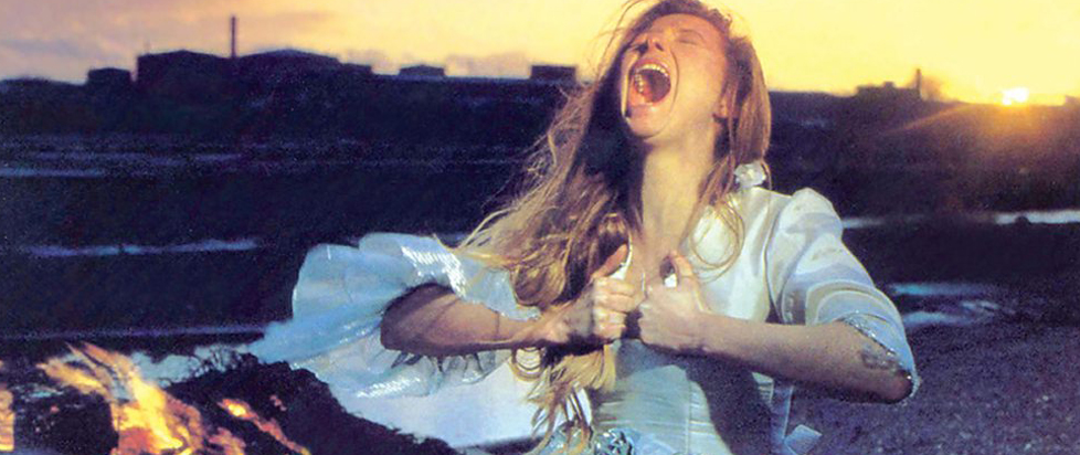 In a still from The Last of England, a woman with long blonde hair wearing a wedding dress rends her dress apart while screaming in front of an open flame.