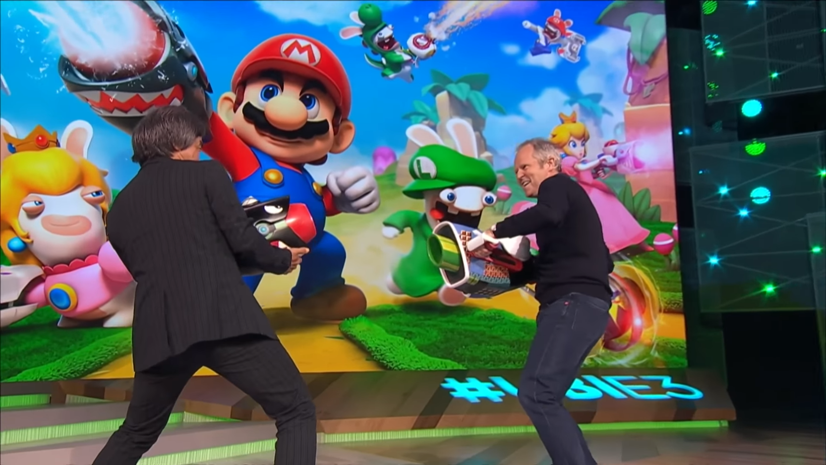 Yves Guillemot and Shigeru Miyamoto onstage at E3, Miyamoto holding an arm cannon that looks like a Bullet Bill pointed at Guillemot, who holds his own oversized blaster, as they stand in front of a screen featuring Mario, Luigi, and the tired looking Rabbids all holding their own firearms