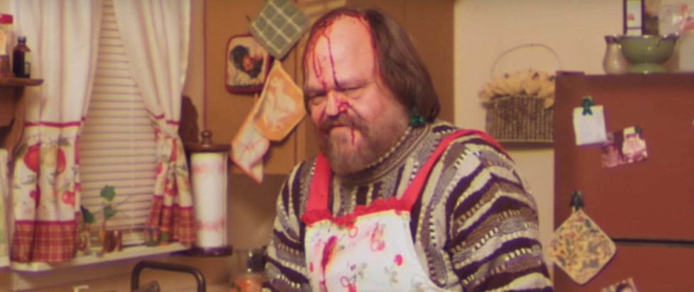 A still from the internet phenomenon "Too Many Cooks" showing the recurring serial killer character in a 1980s kitchen with a bloodied face and apron.