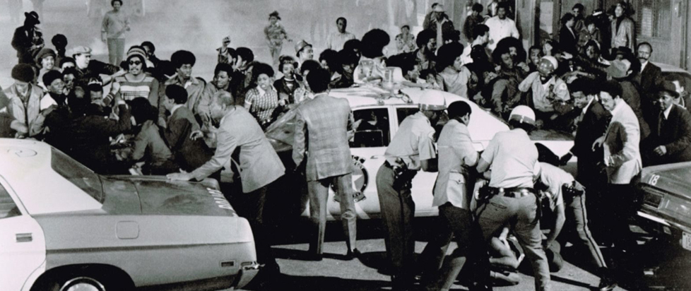 A black and white photograph of several Black Americans rioting in the street in the 1970s.